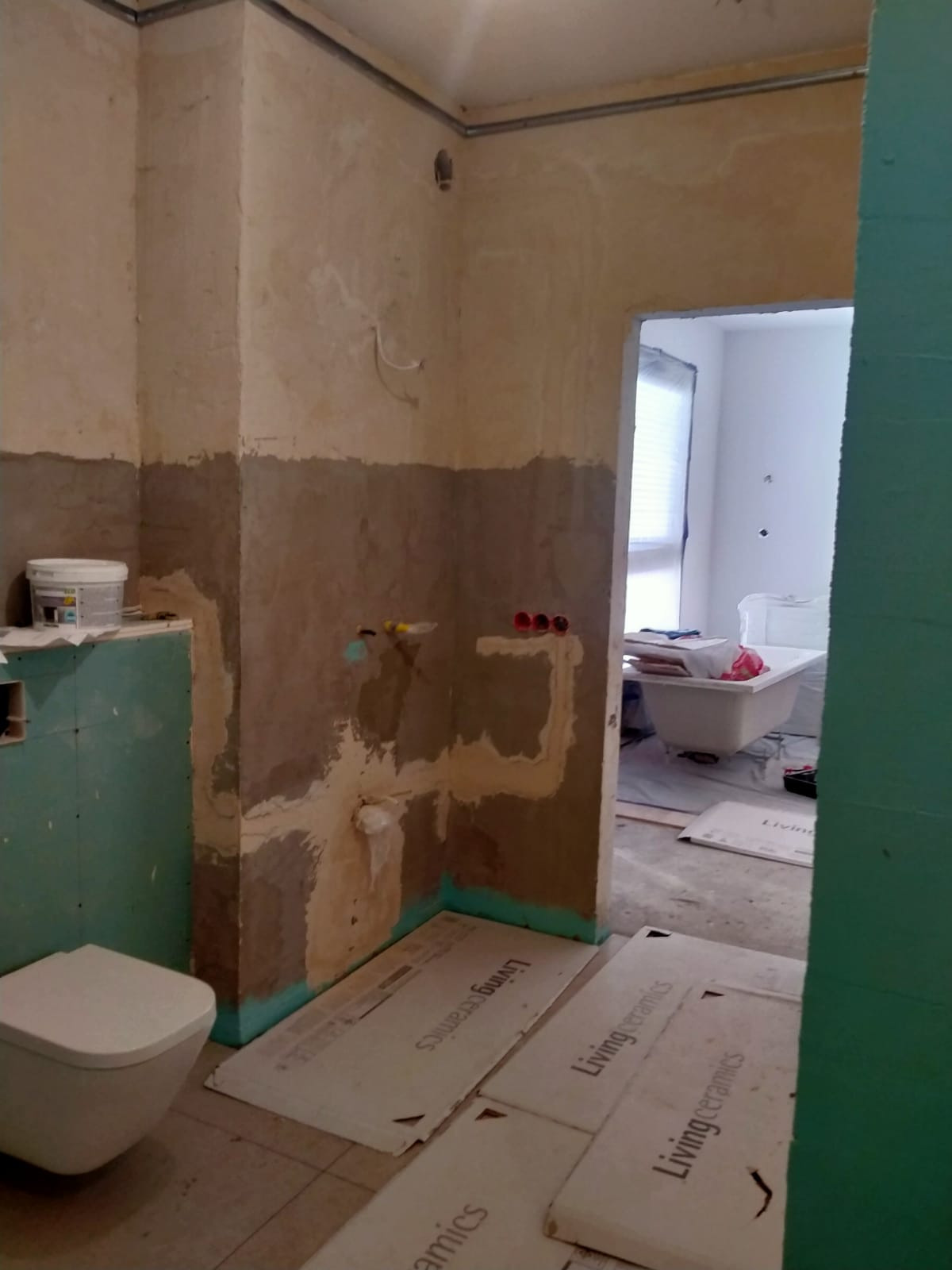 Łazienka w trakcie remontu: ściany częściowo przygotowane pod płytki, widoczne instalacje, na podłodze kartony z nazwą Living Ceramics, toaleta zamontowana, w tle wanna.