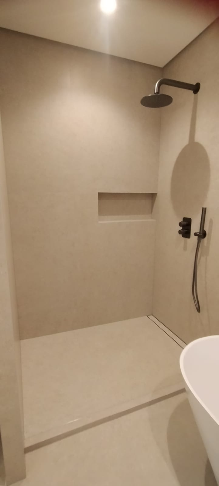 Nowoczesna kabina prysznicowa walk-in z beżowymi płytkami, czarną armaturą i odpływem liniowym, oświetlona górnym światłem.