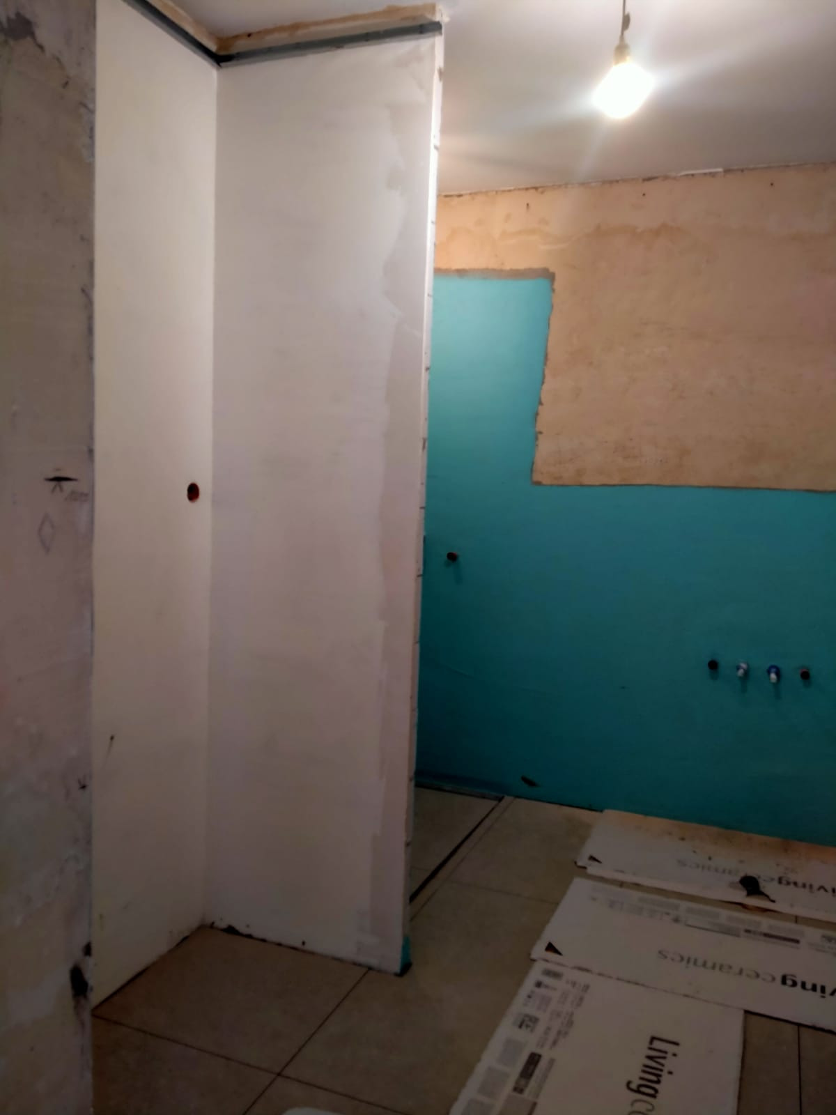 Niedokończona łazienka w trakcie remontu: ściany częściowo pokryte tynkiem, fragment ściany pomalowany na turkusowo, widoczne rury, na podłodze płyty ceramiczne Living Ceramics.