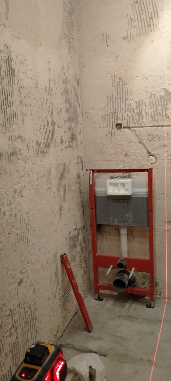 Stelaż podtynkowy WC marki TECE zamontowany w rogu łazienki, widoczny laser poziomujący, ściany przygotowane pod płytki, poziomica oparta o ścianę.