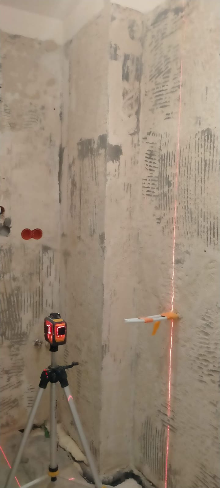 Przygotowanie ściany pod płytki w rogu pomieszczenia, widoczny laser krzyżowy na statywie wyznaczający pion, ściana z resztkami kleju po starych płytkach, dwa otwory pod gniazdka elektryczne.