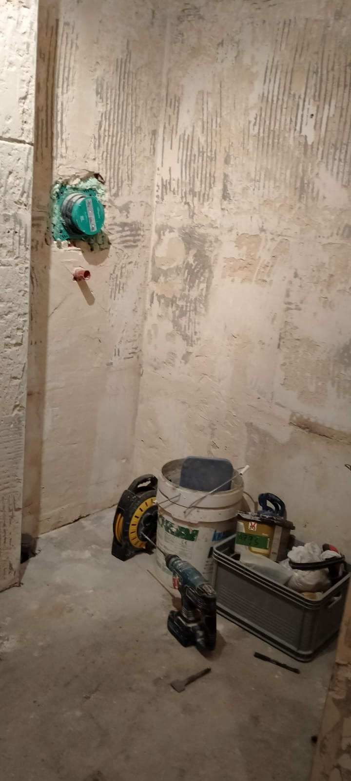 Przygotowanie do remontu łazienki: ściany z usuniętymi płytkami, odsłonięta instalacja wodna z zielonym zaworem, wiadro z zaprawą, wiertarka akumulatorowa i przedłużacz na podłodze.