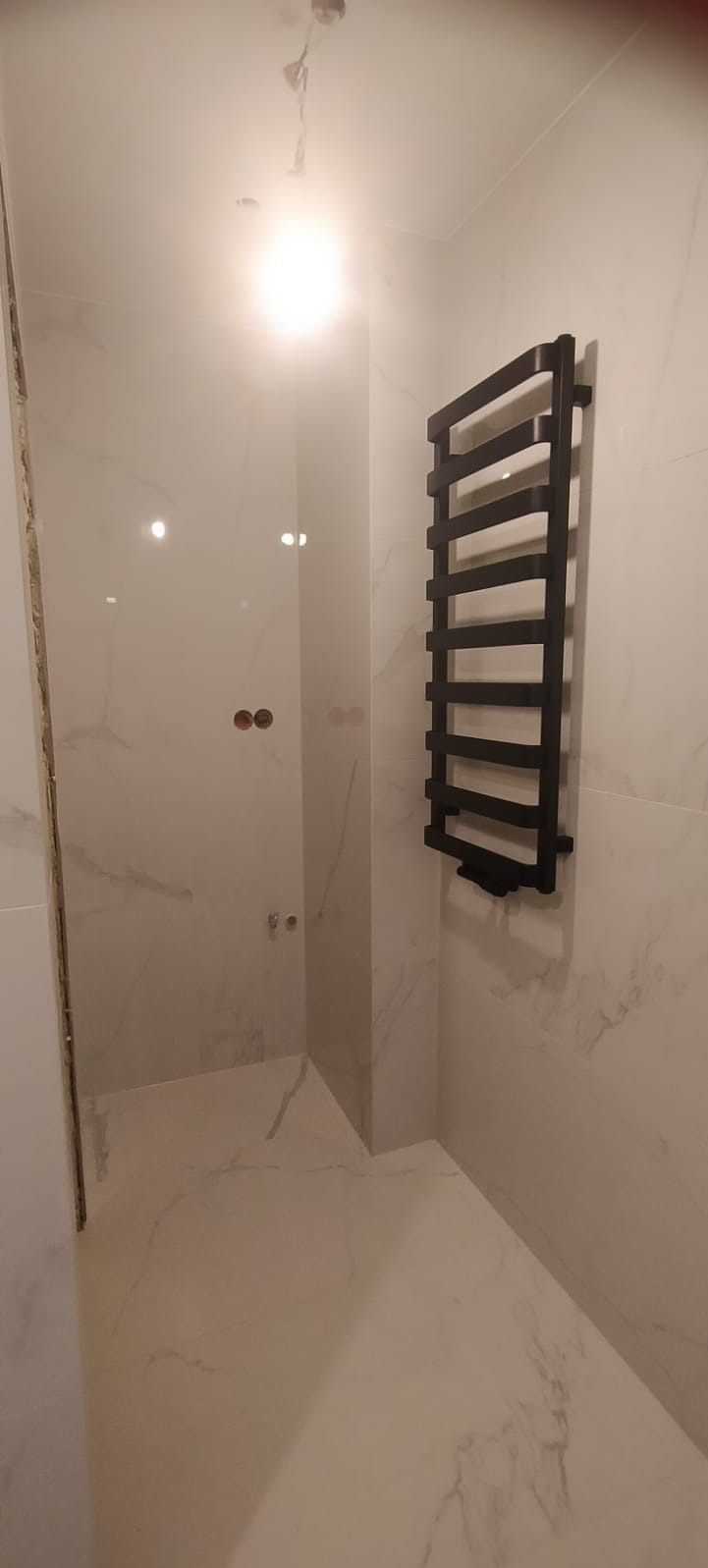 Wykończona łazienka z białymi płytkami imitującymi marmur i czarnym grzejnikiem drabinkowym, widoczne puszki elektryczne bez osprzętu.