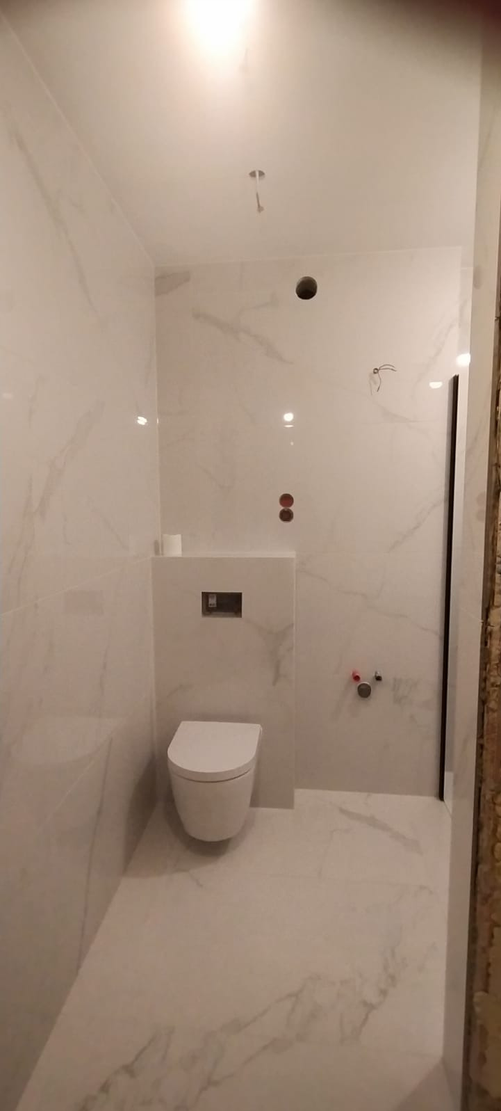 Nowoczesna, biała łazienka z wiszącą toaletą i płytkami imitującymi marmur, widoczne instalacje wodne i otwory w suficie przed montażem oświetlenia.