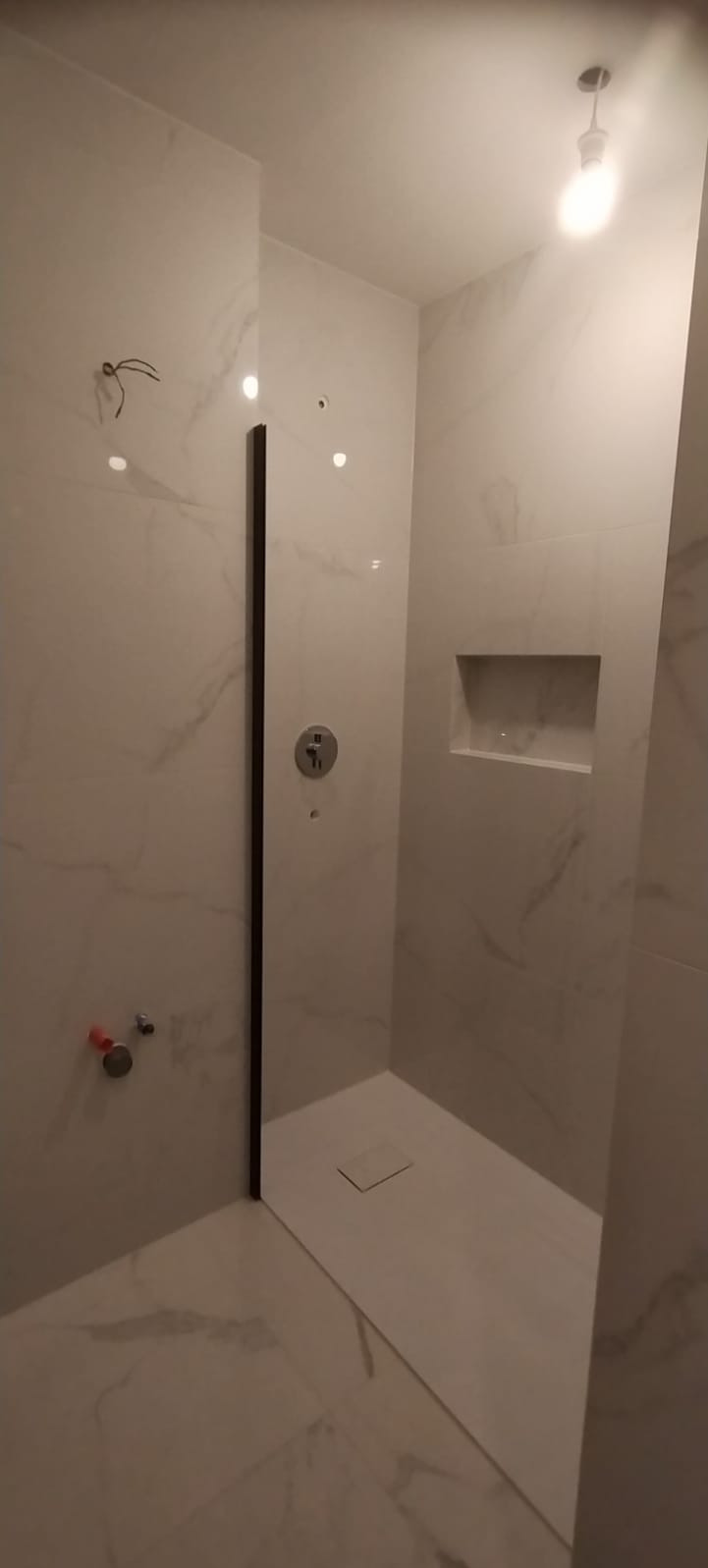 Nowoczesna kabina prysznicowa z marmurowymi płytkami, szklaną ścianką z ciemną ramą i wnęką w ścianie, widoczne przyłącza wodne.