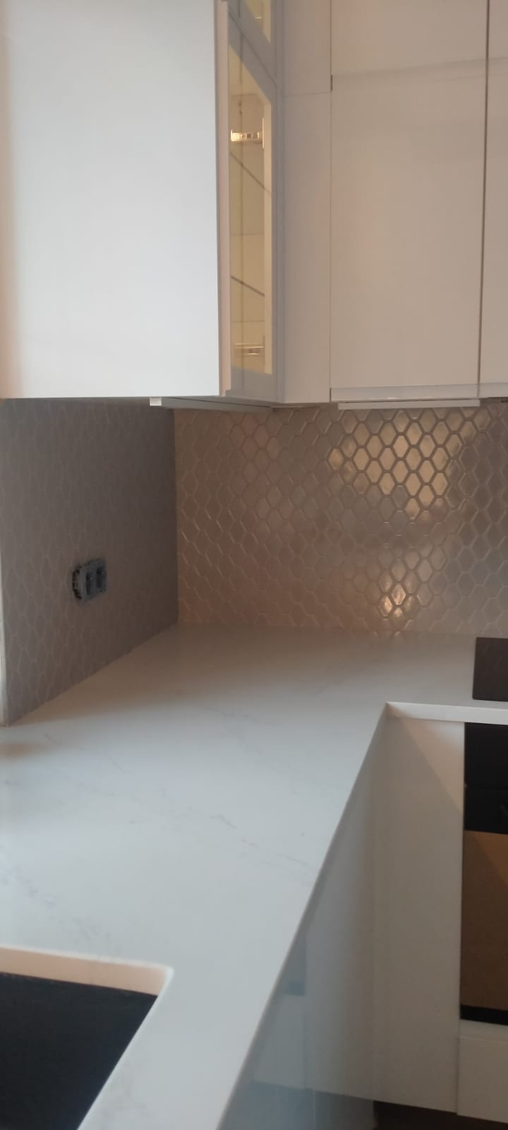 Ukończona kuchnia z białymi szafkami i mozaiką w odcieniach szarości na ścianie, widoczne gniazdko elektryczne.