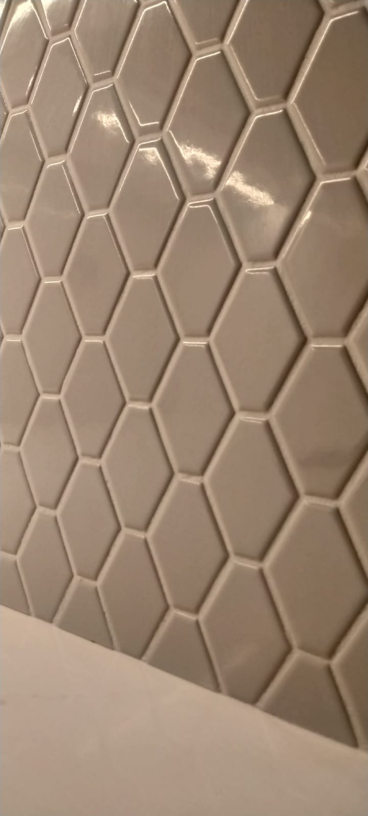 Beżowe, błyszczące, heksagonalne płytki ceramiczne ułożone na ścianie, widoczne z bliska, z fragmentem białej powierzchni w dolnej części kadru.
