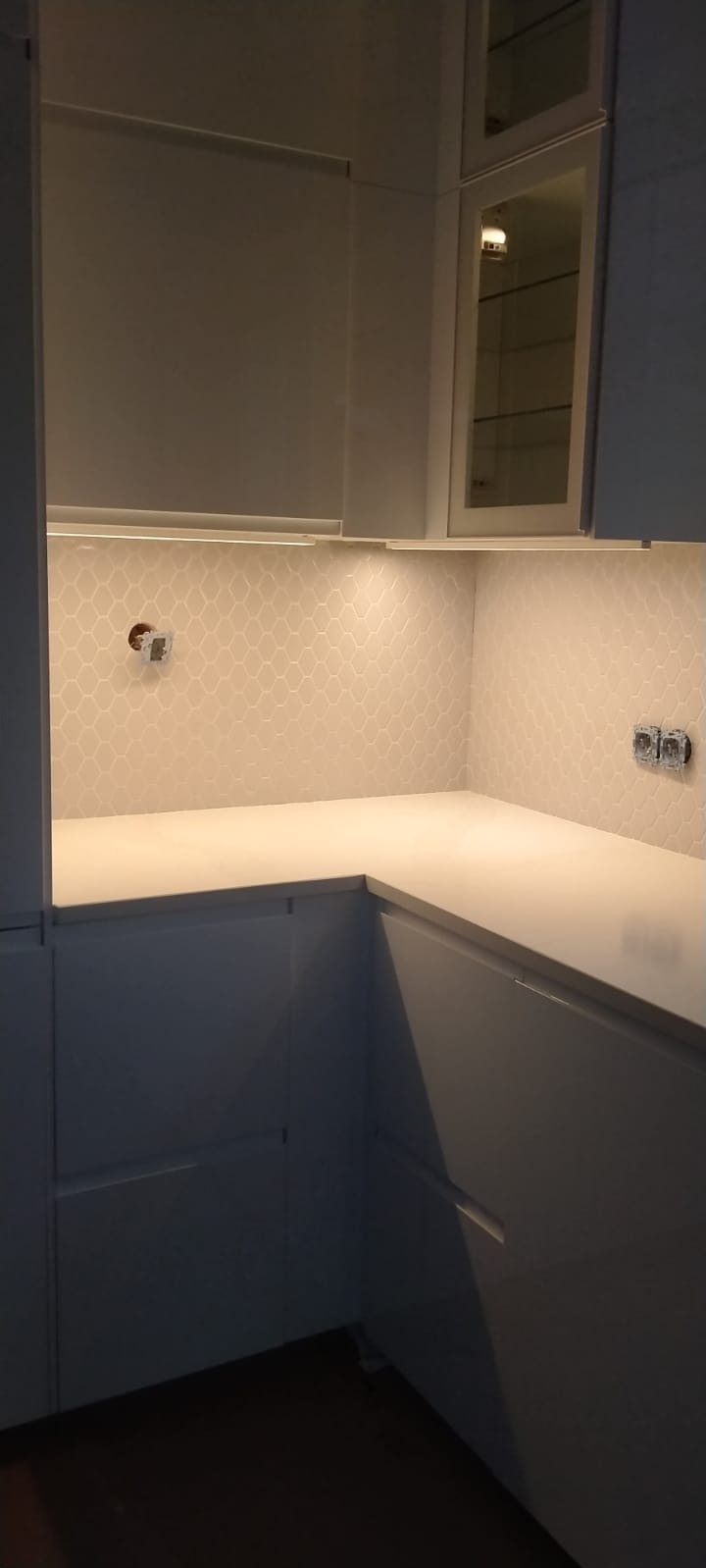 Fragment kuchni z białymi szafkami i blatem, podświetlana mozaika na ścianie, widoczne gniazdka elektryczne i otwór na instalację.