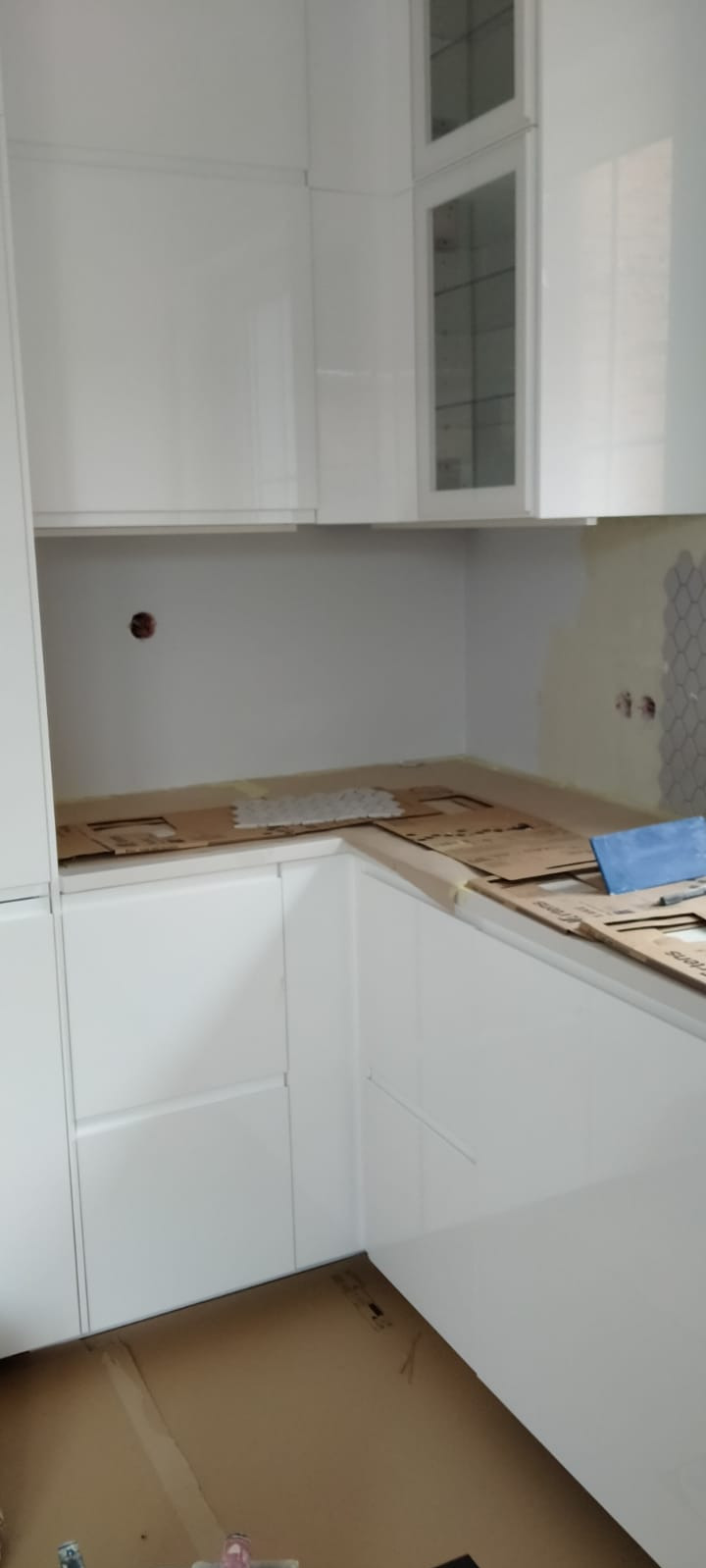 Biała zabudowa kuchenna w trakcie prac wykończeniowych: częściowo ułożona mozaika na ścianie, blat zabezpieczony kartonem, widoczne ślady po wierceniu w ścianie.