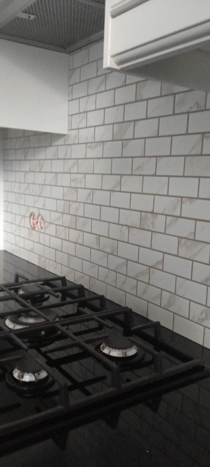 Białe, marmurkowe płytki ułożone na ścianie nad czarną płytą gazową, widoczne otwory na gniazdka elektryczne.