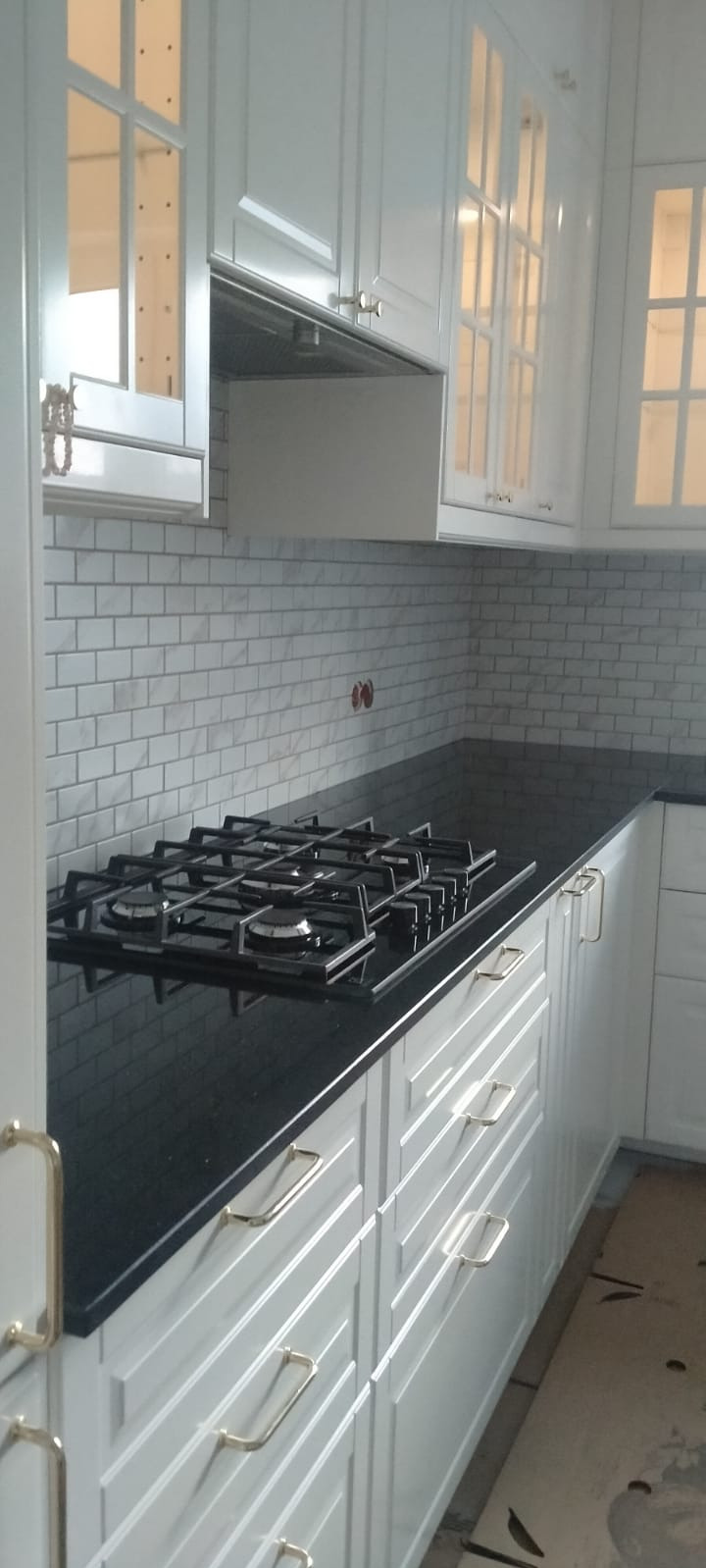 Nowoczesna kuchnia z białymi szafkami z frezowanymi frontami i złotymi uchwytami, czarny blat z zamontowaną płytą gazową oraz białe płytki imitujące cegłę na ścianie.