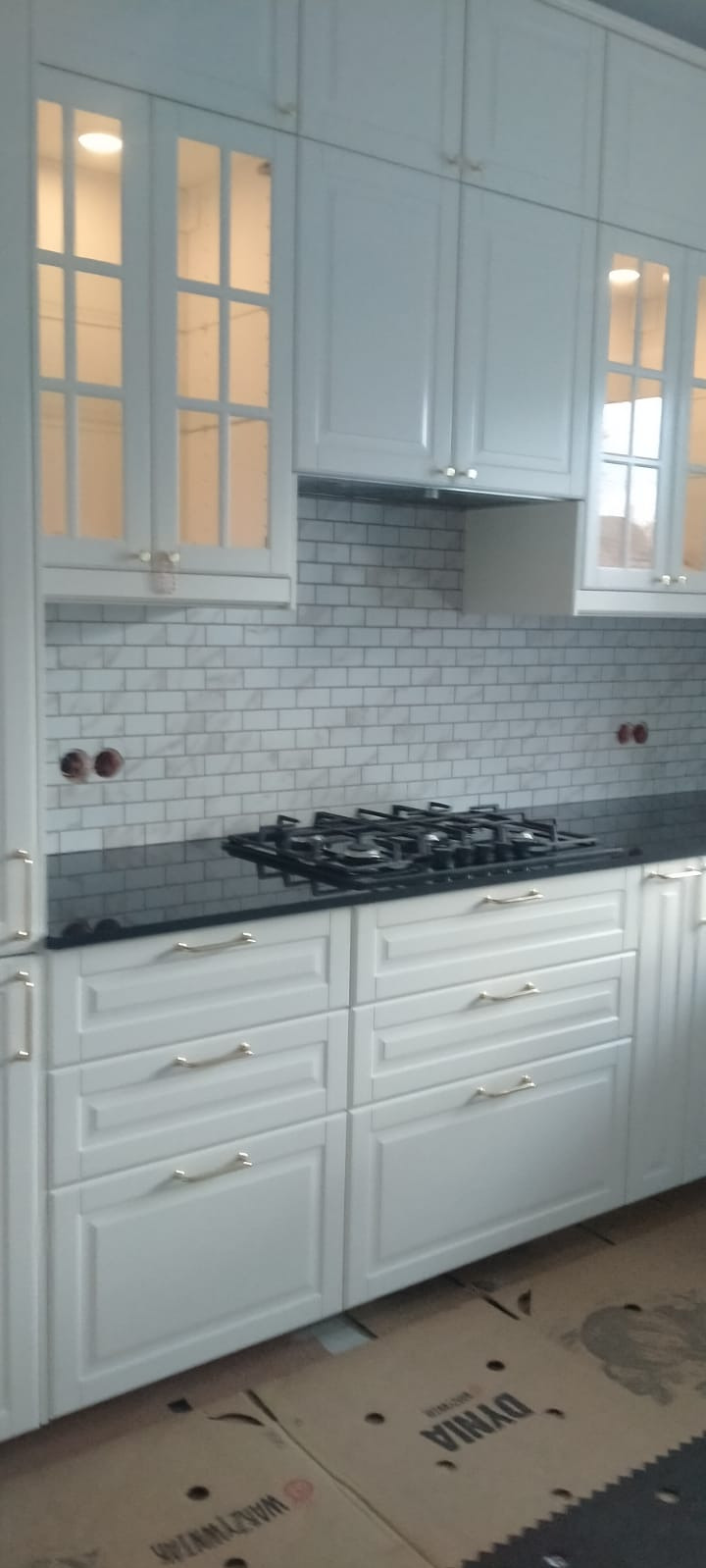 Biała zabudowa kuchenna z dolnymi szafkami z szufladami i górnymi szafkami z przeszkleniami, czarny blat i białe płytki na ścianie, widoczne dwa otwory na ścianie.