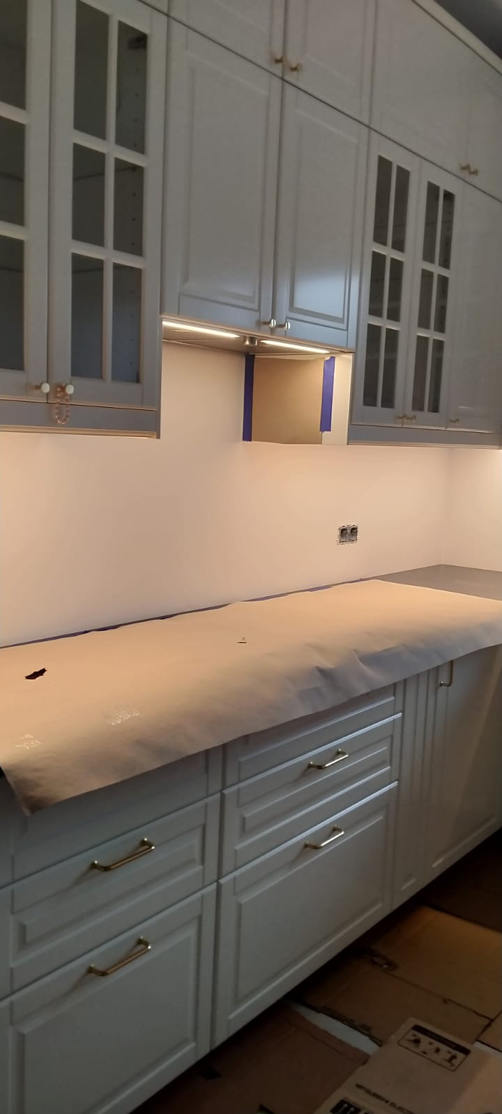 Białe szafki kuchenne z przeszklonymi frontami, zabezpieczone papierem malarskim przed malowaniem ścian, widoczne gniazdko elektryczne i taśma malarska.