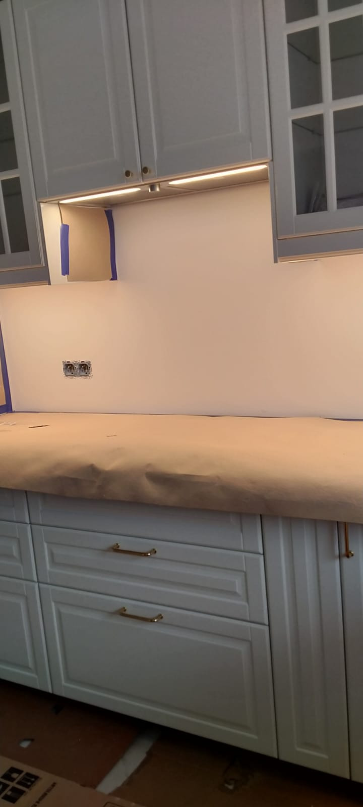 Przygotowanie do malowania ściany w kuchni: zabezpieczony blat papierem maskującym, ściana oklejona taśmą malarską, widoczne gniazdka elektryczne.