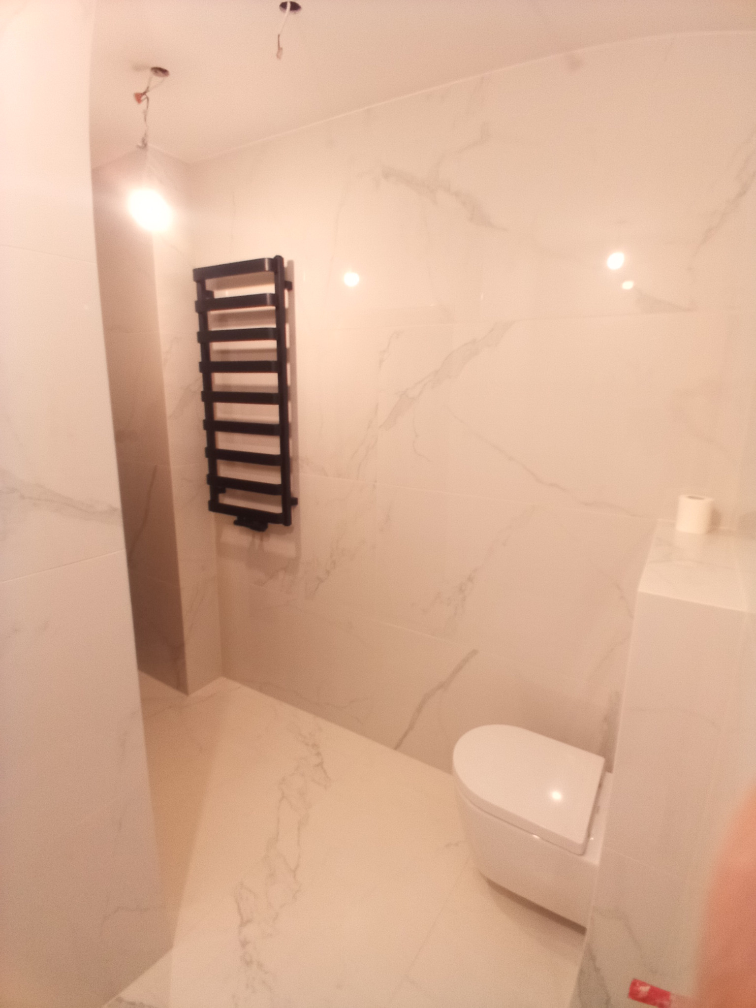 Wykończona łazienka w jasnych płytkach imitujących marmur, z podwieszaną toaletą i czarnym grzejnikiem drabinkowym. Widoczne wystające kable elektryczne ze ściany.