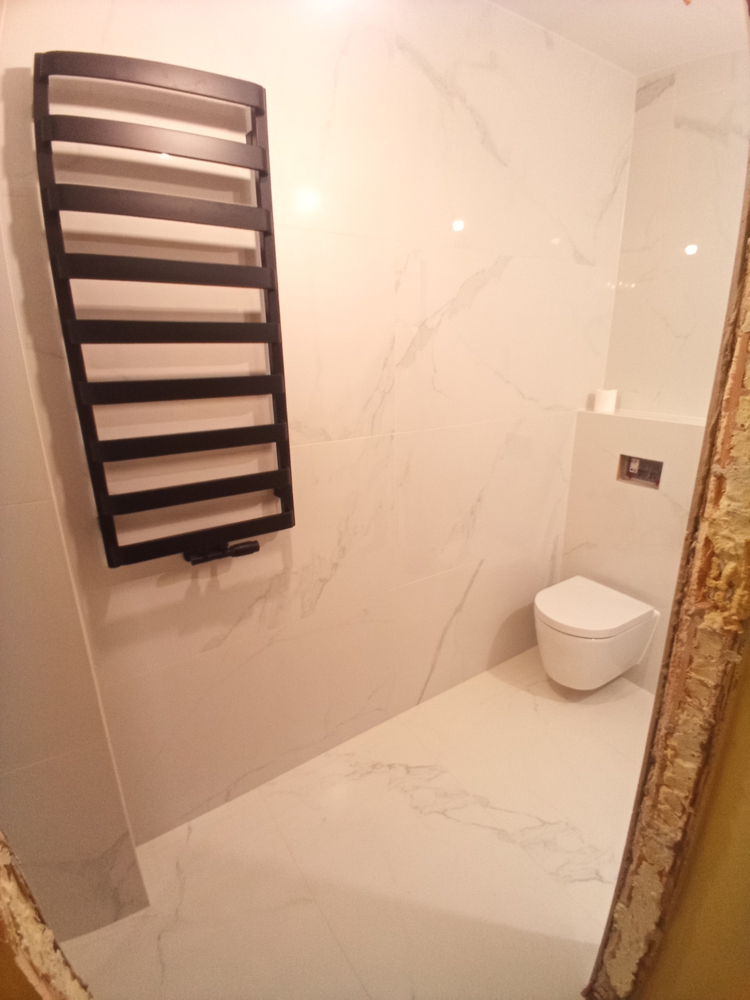 Nowoczesna łazienka z białymi płytkami imitującymi marmur, wiszącą toaletą i czarnym grzejnikiem drabinkowym, widok przez futrynę w trakcie remontu.