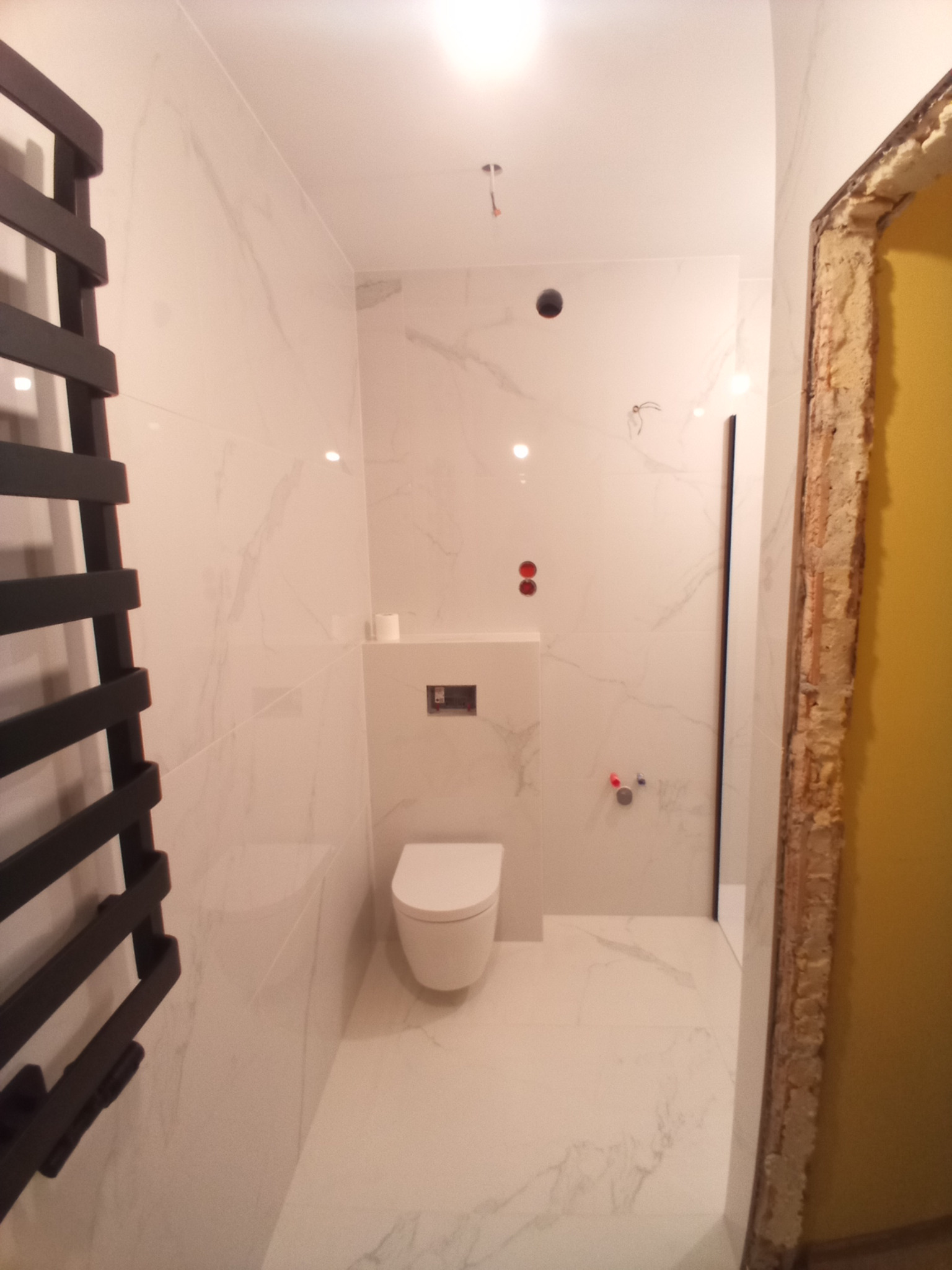 Nowoczesna łazienka w trakcie remontu: białe marmurowe płytki, podwieszana toaleta, grzejnik drabinkowy, widoczne przygotowania do montażu ościeżnicy.