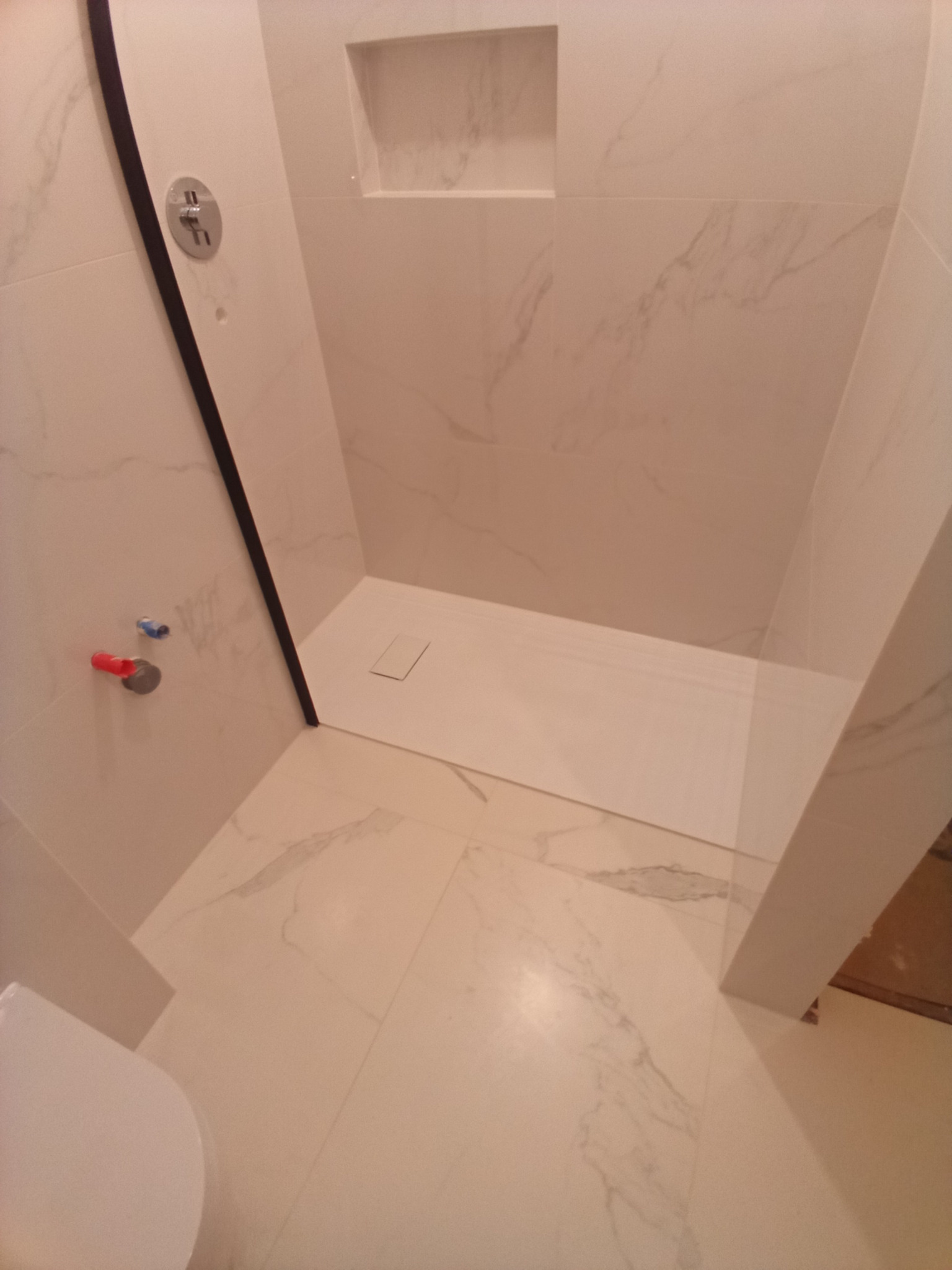 Wykończona kabina prysznicowa z białymi płytkami imitującymi marmur, widoczne elementy armatury i odpływ liniowy.