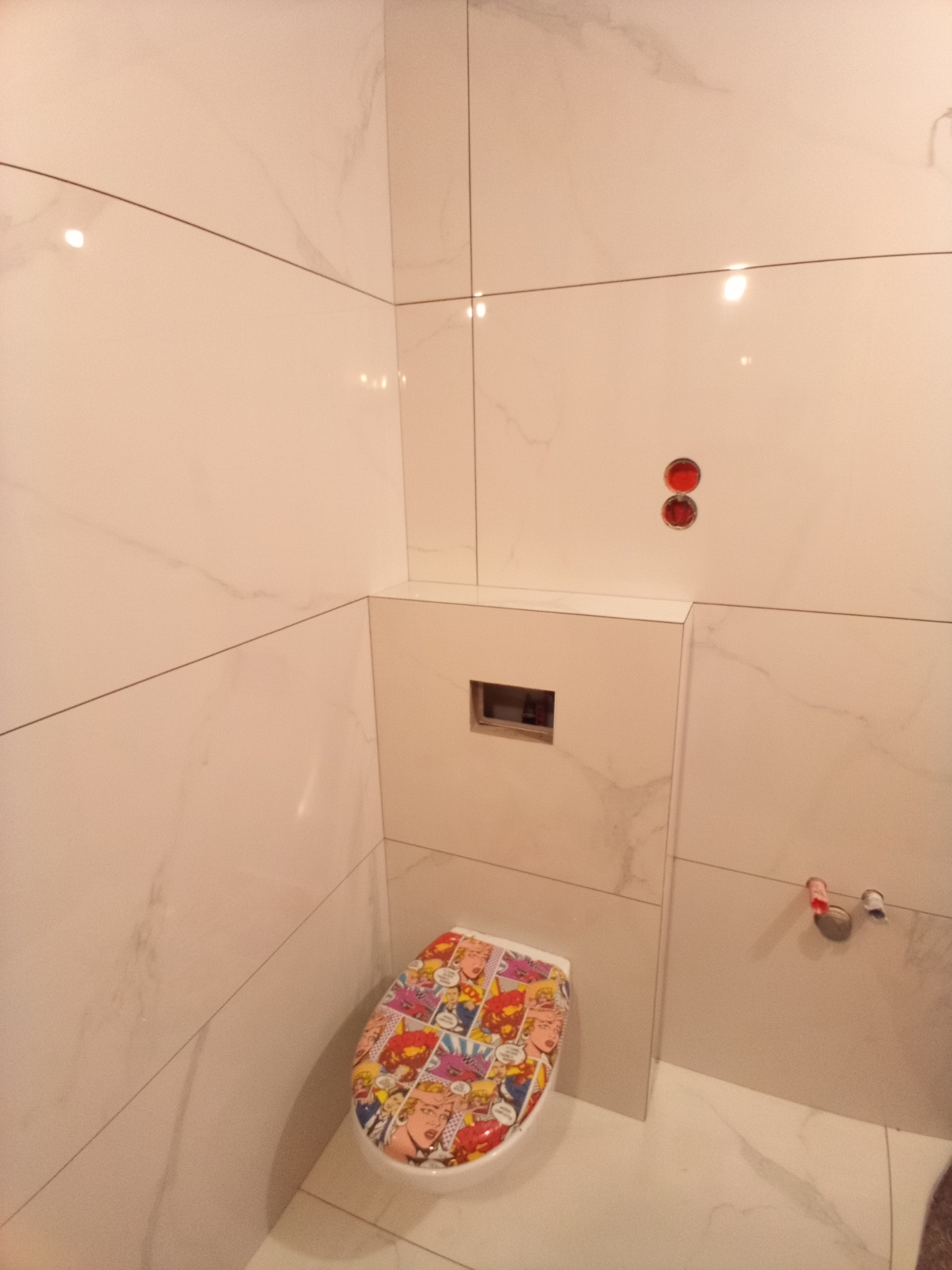 Wykończona łazienka z białymi, marmurkowymi płytkami na ścianach i podłodze, z toaletą z zabudowaną spłuczką i deską klozetową z motywem komiksowym.
