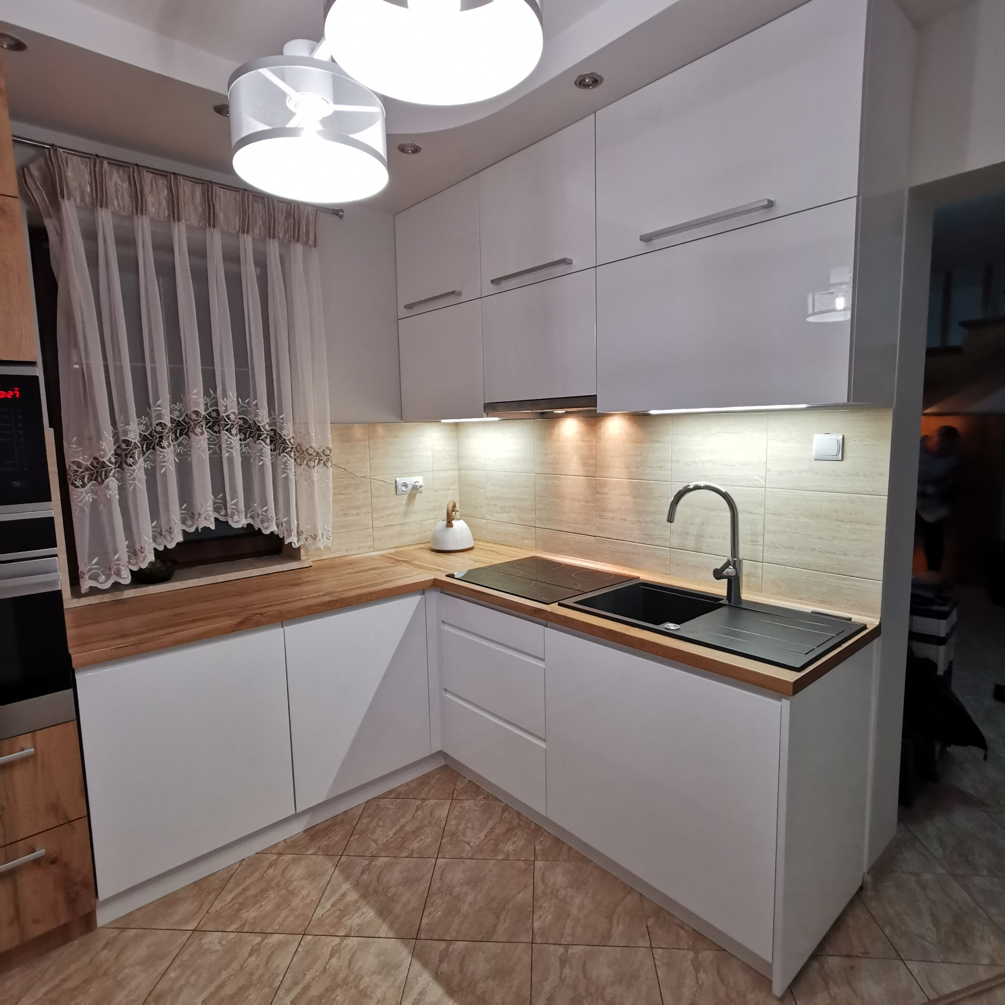 Nowoczesna kuchnia z białymi szafkami i drewnianym blatem, czarny zlew i płyta indukcyjna, beżowe płytki na ścianie, oświetlenie podszafkowe i firanka w oknie.