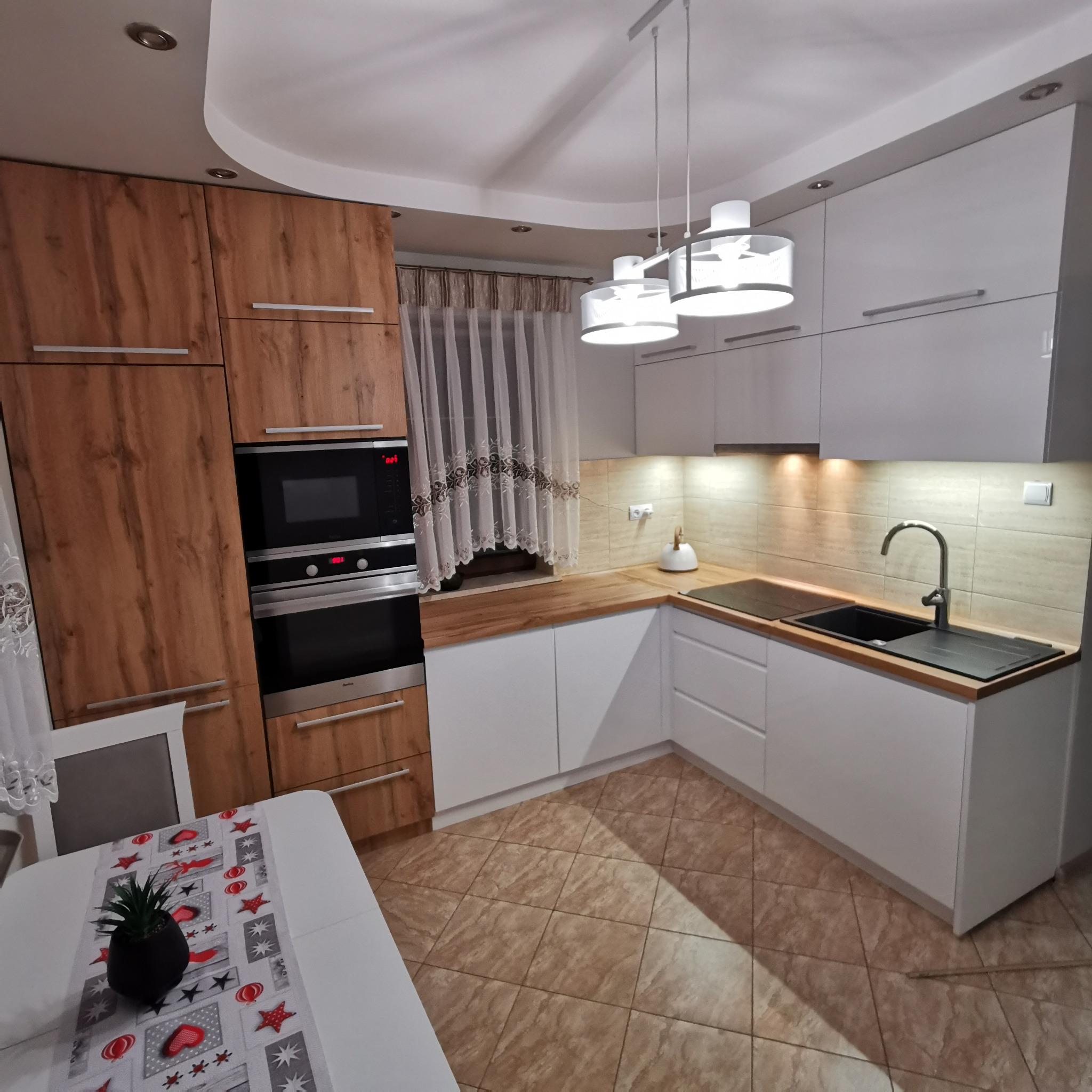 Nowoczesna kuchnia z frontami w kolorze drewna i białymi szafkami, oświetlona trzema lampami wiszącymi, z czarnym zlewem i płytą indukcyjną, widoczny stół z dekoracyjnym bieżnikiem i rośliną...