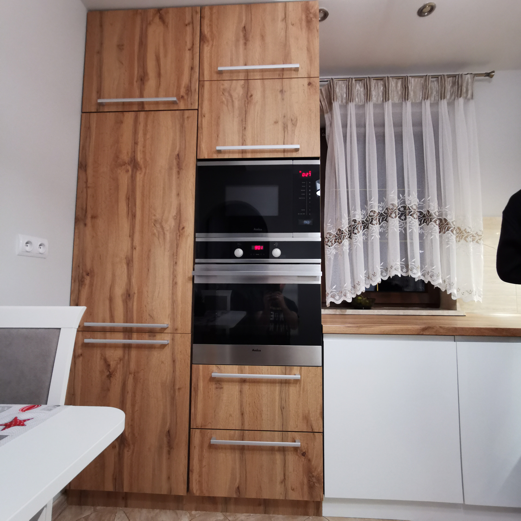 Nowoczesna zabudowa kuchenna z fornirem drewnopodobnym, wbudowanym piekarnikiem i mikrofalówką Amica, białe dolne szafki, firanka w oknie.