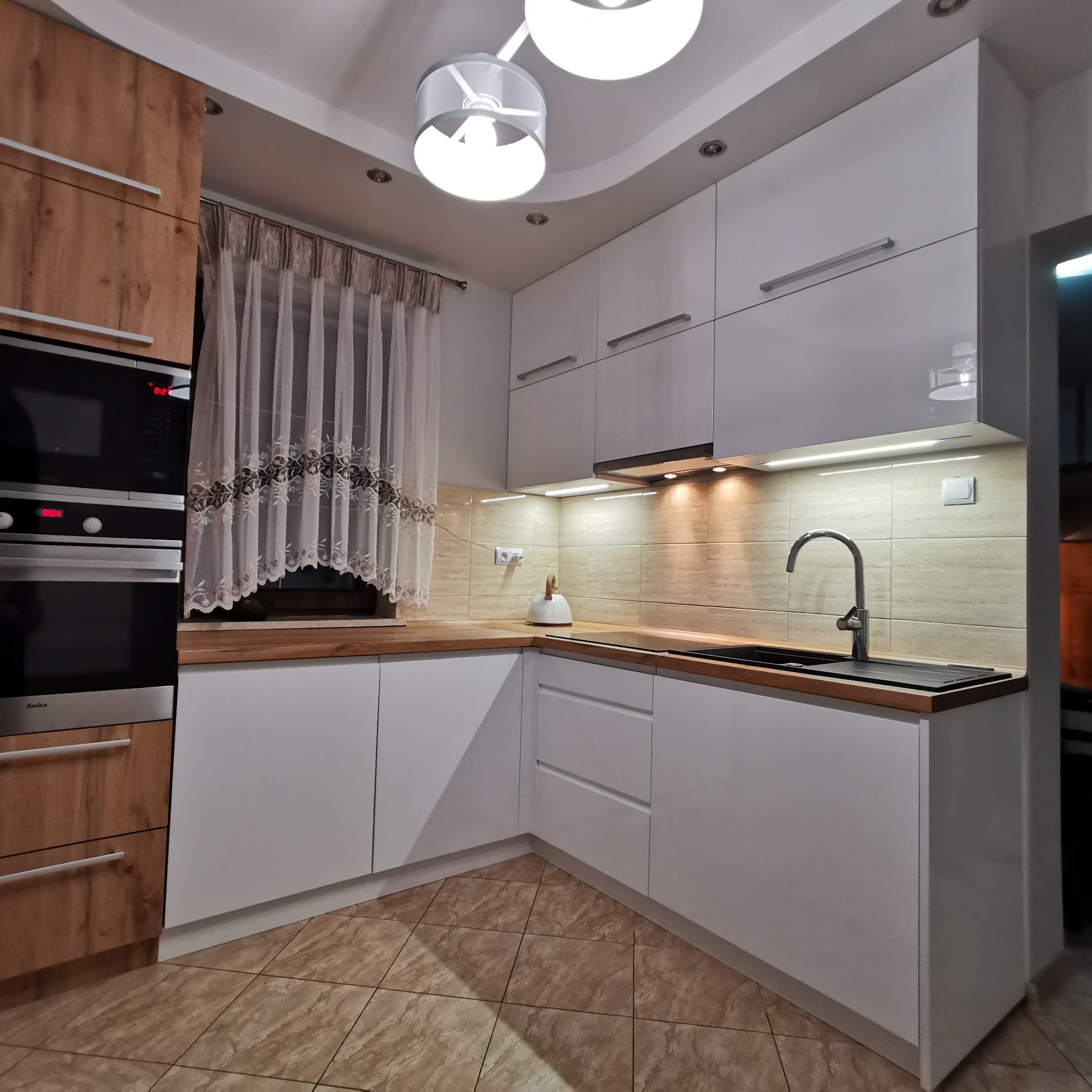 Nowoczesna kuchnia w Malborku z białymi frontami i drewnianym blatem, oświetlona lampami sufitowymi i listwami LED, z widocznym piekarnikiem i zasłoną w oknie.