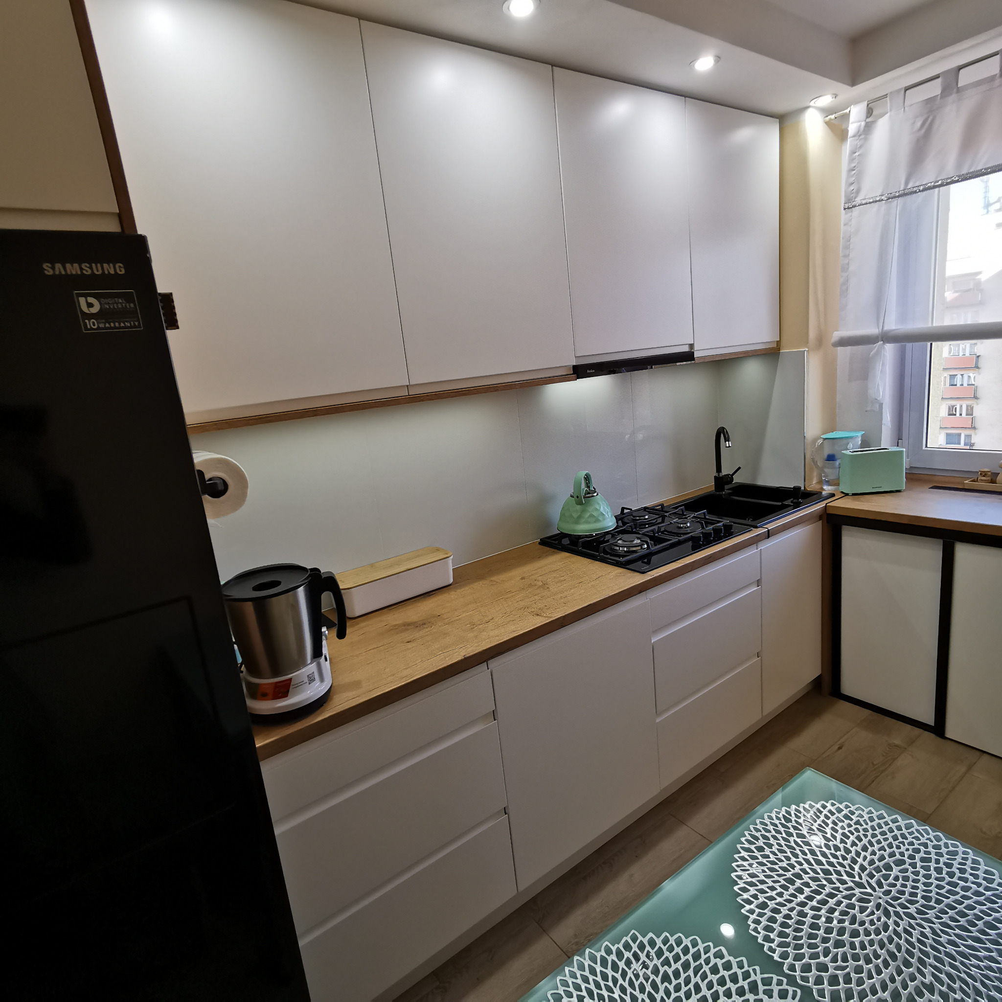 Jasna kuchnia z białymi szafkami i drewnianym blatem, czarny zlew i kuchenka gazowa, pastelowy czajnik i toster, nowoczesny design.