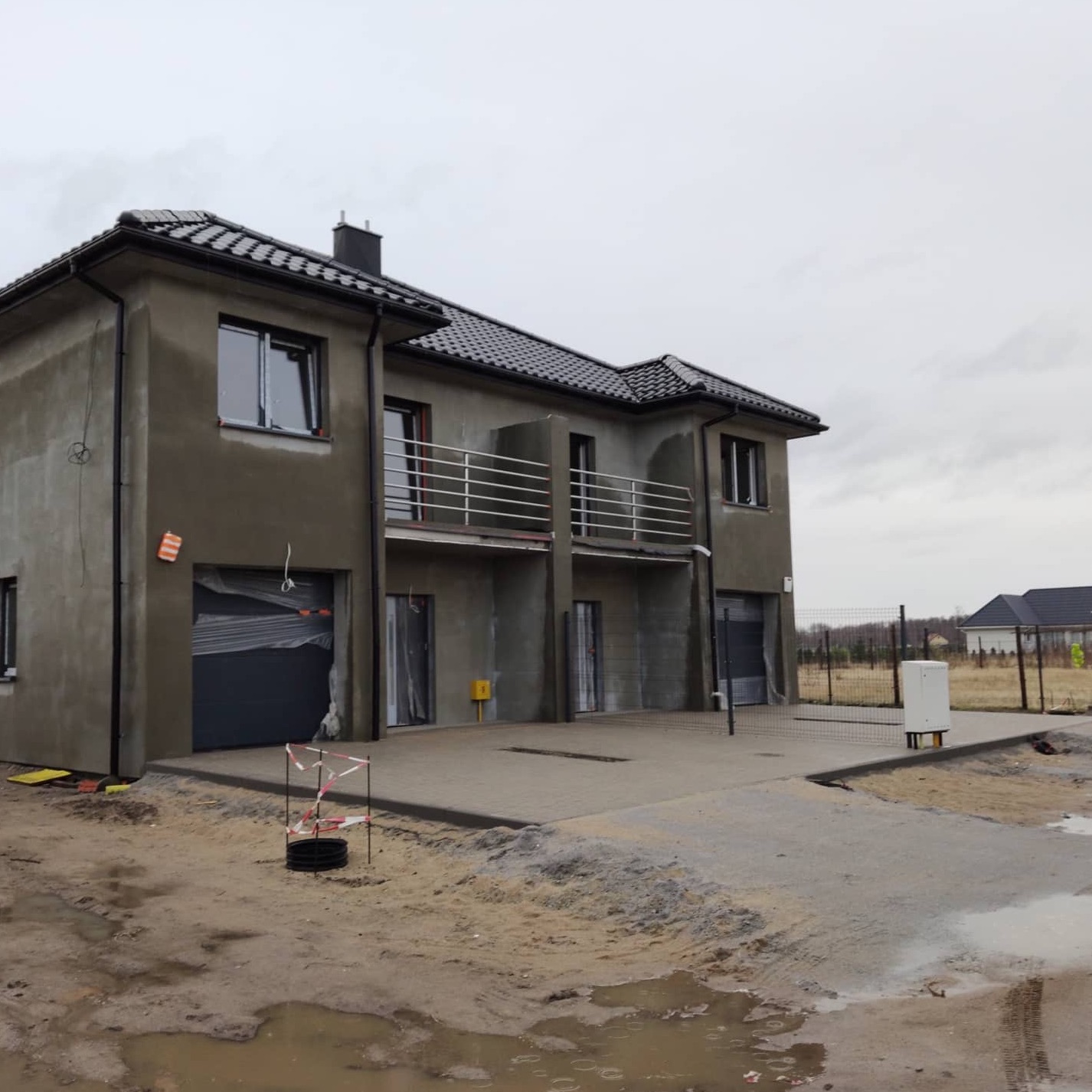Surowy stan bliźniaka z szarym tynkiem zewnętrznym, czarną dachówką i garażami zabezpieczonymi folią, widoczne ślady prac budowlanych na zewnątrz.