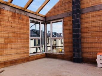 Wnętrze domu w trakcie budowy: ściany z bloczków ceramicznych, dwa okna narożne z białymi ramami, widoczne elementy konstrukcji dachu oraz komin z szarych pustaków.