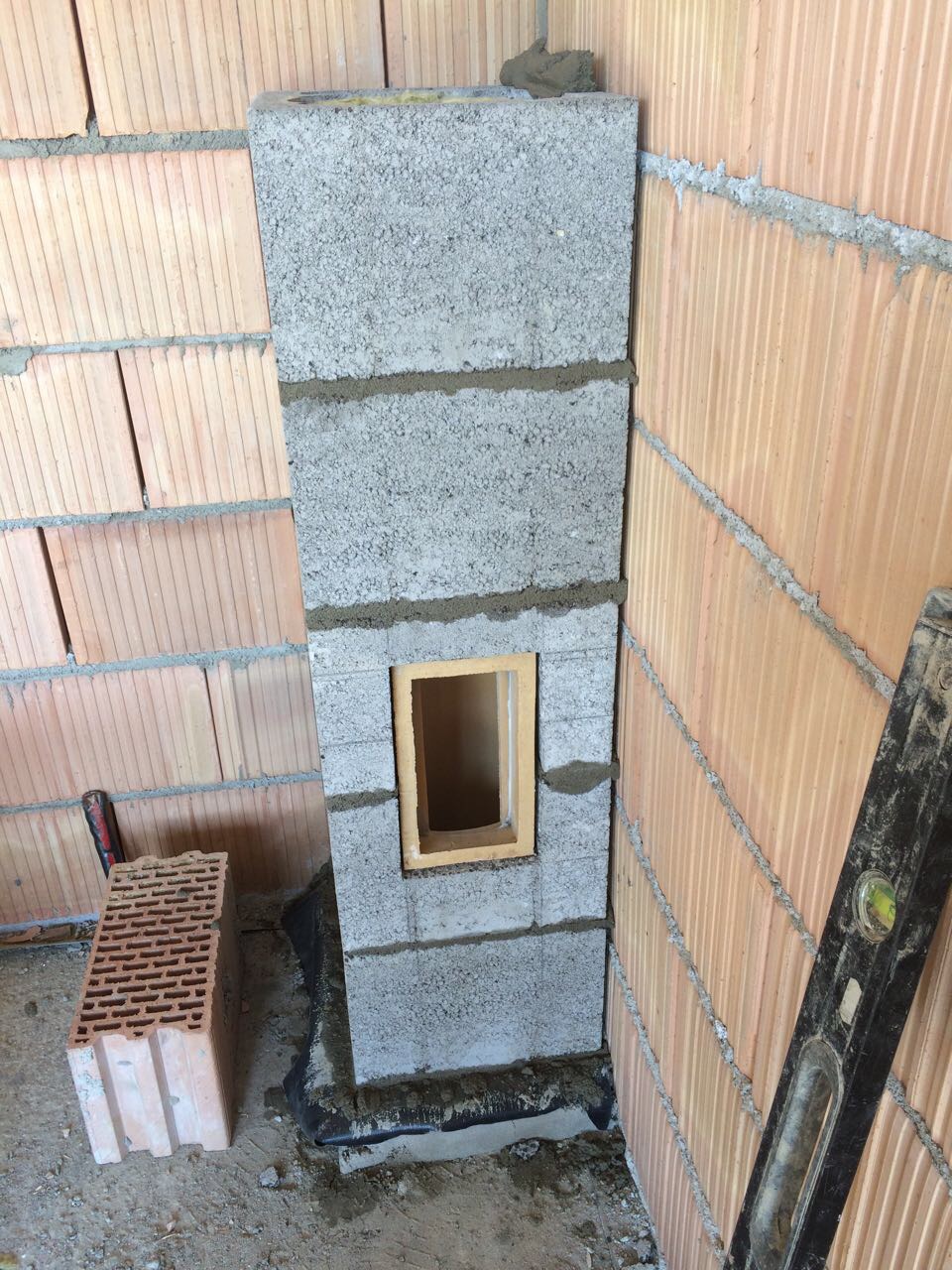 Wznoszenie komina z bloczków betonowych wewnątrz budynku, widoczne bloczki, zaprawa, fragment ściany z pustaków ceramicznych oraz poziomica.