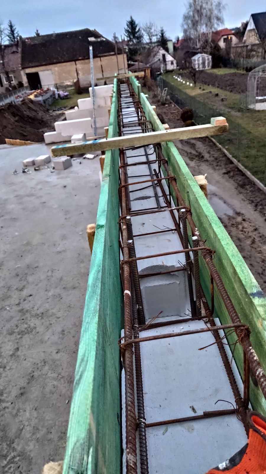 Wiązanie zbrojenia w szalunku fundamentowym z bloczków betonowych i drewnianych desek, widok z góry na plac budowy w pochmurny dzień.