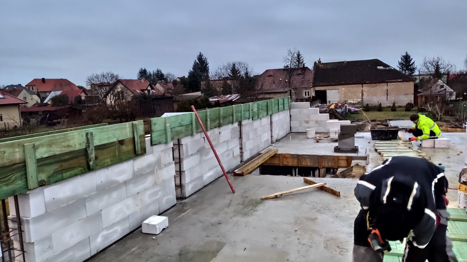 Wznoszenie ścian z bloczków betonowych na fundamencie, widoczne szalunki i zbrojenie, w tle zabudowa jednorodzinna w pochmurny dzień.