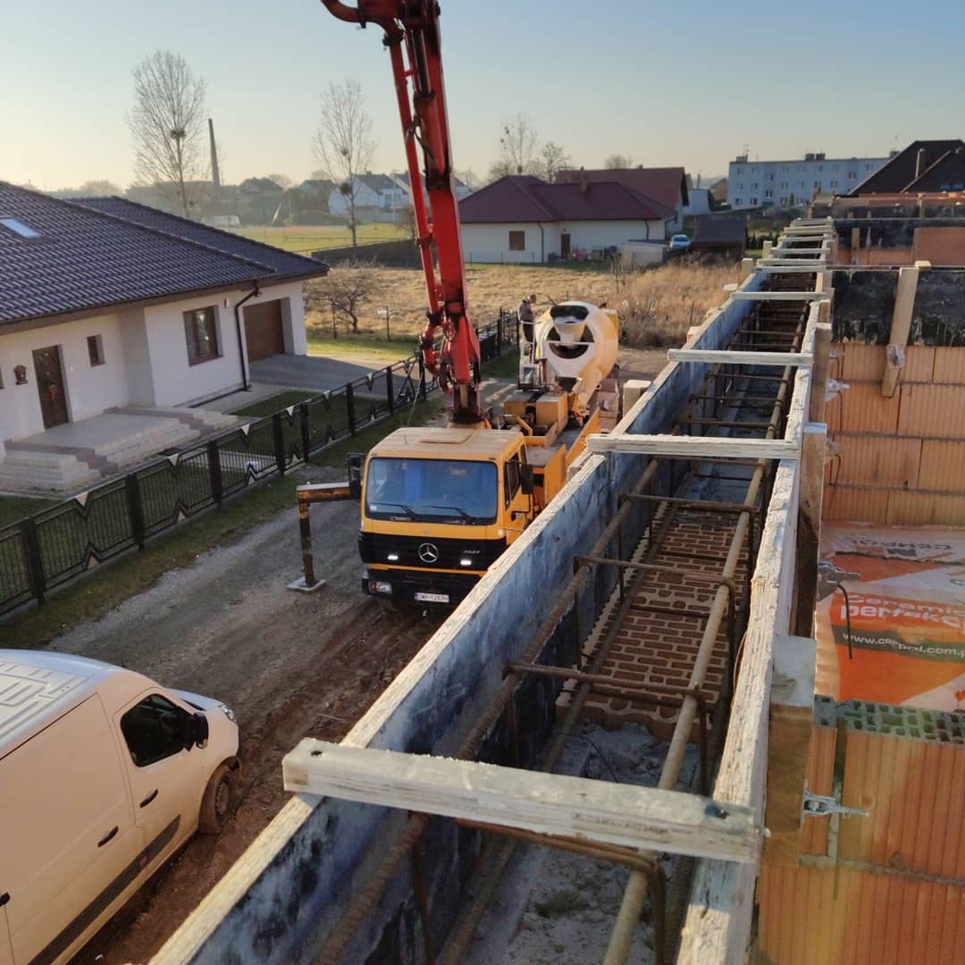 Wylewanie betonu do szalunków zbrojonych stalą na budowie domu jednorodzinnego, widoczny mikser do betonu z pompą oraz fragment muru z pustaków ceramicznych.
