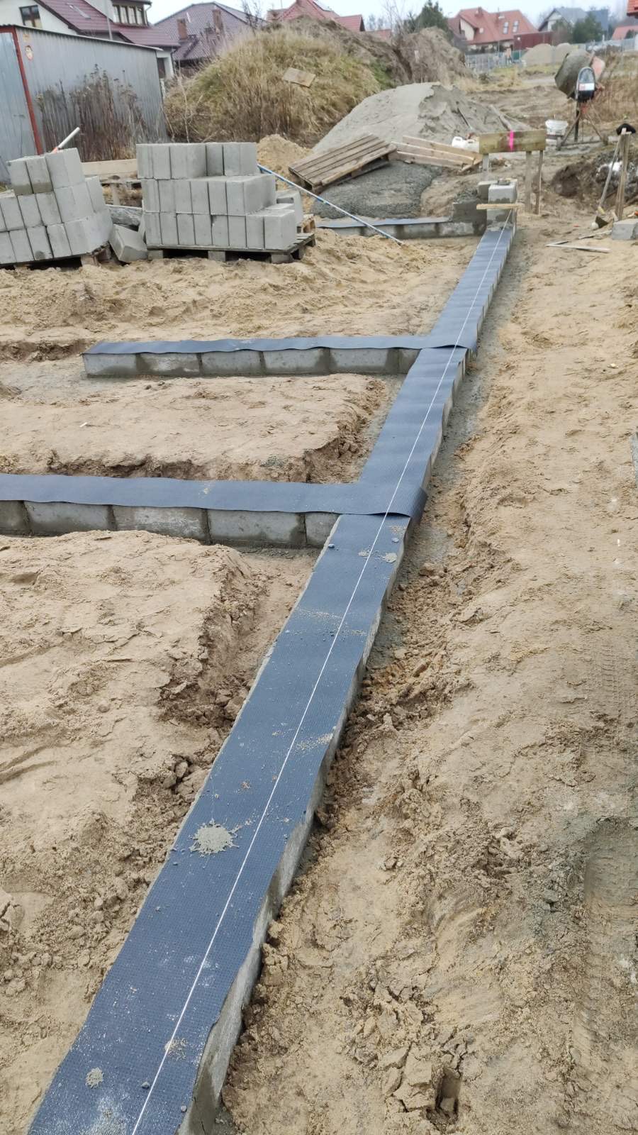 Wykopane fundamenty z ułożonymi bloczkami betonowymi, zabezpieczone czarną folią, wytyczone sznurkiem, w tle hałda piasku i częściowo zabudowane tereny.
