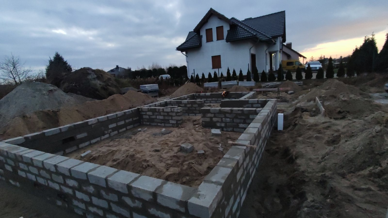 Wznoszenie fundamentów z bloczków betonowych na piaszczystym podłożu pod budowę domu jednorodzinnego w tle, widoczne hałdy piasku i materiałów budowlanych.