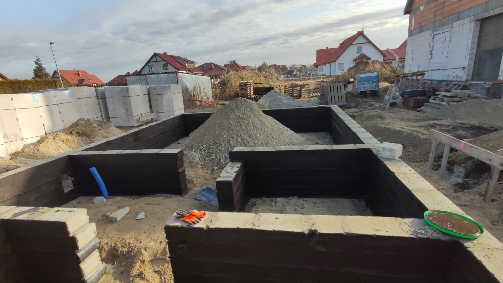 Wykopane fundamenty pod dom, zabezpieczone hydroizolacją, z widoczną stertą żwiru, bloczkami betonowymi w tle i elementami instalacji. Widoczny krajobraz podmiejski z domami w tle.