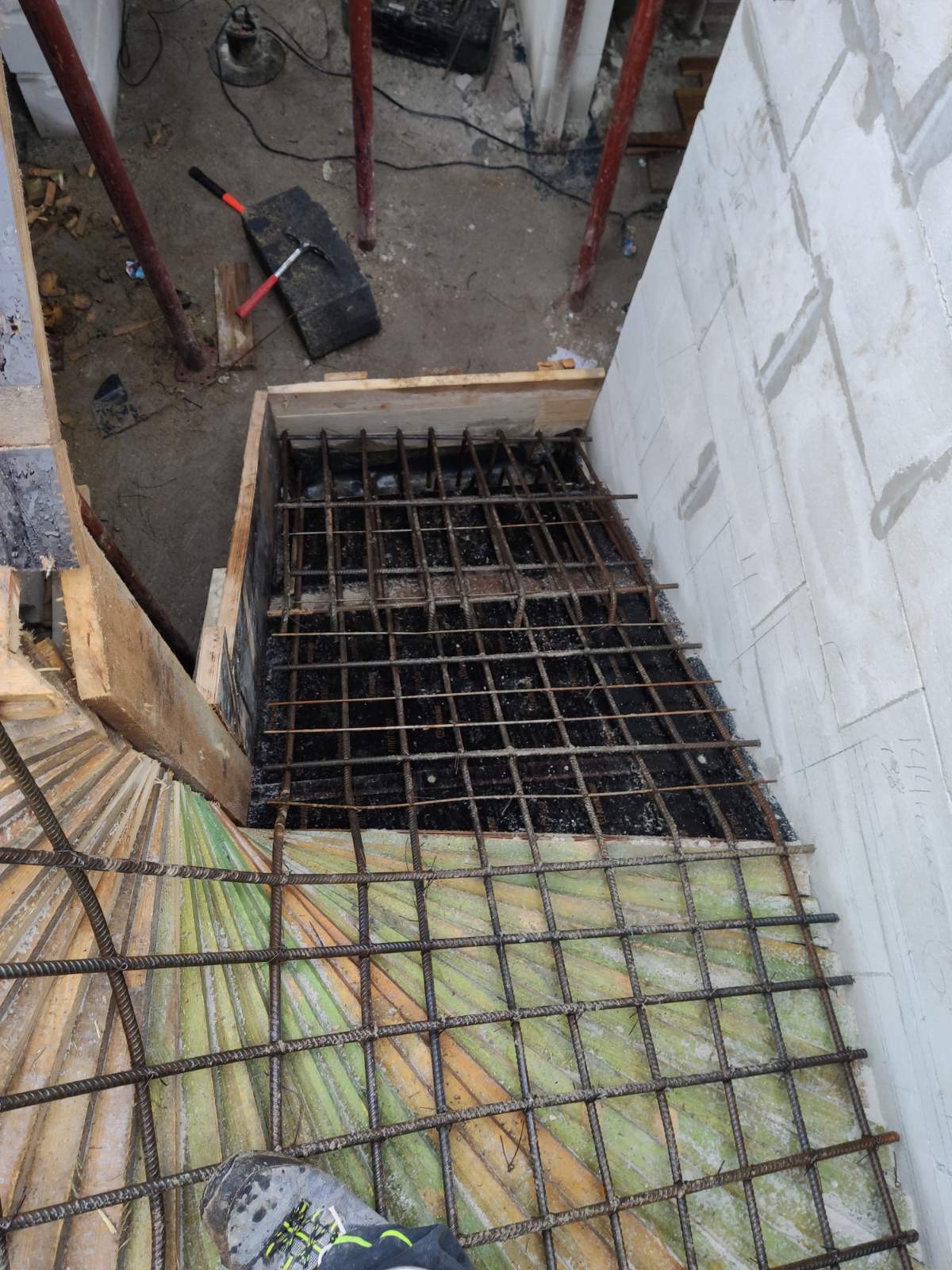 Zbrojenie fundamentu w drewnianym szalunku, widok z góry na siatkę zbrojeniową, beton i fragment ściany z bloczków. Na pierwszym planie fragment buta pracownika.