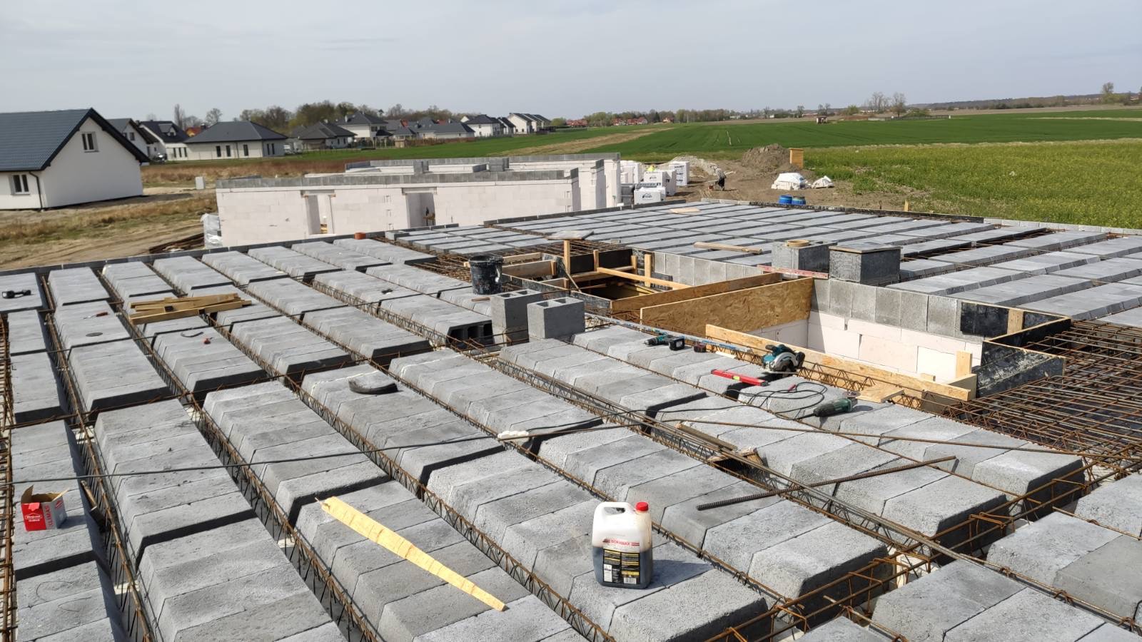 Wiązanie zbrojenia i układanie betonowych bloczków stropowych na budowie domu jednorodzinnego, w tle widoczne kolejne budynki w stanie surowym otwartym i pola uprawne.