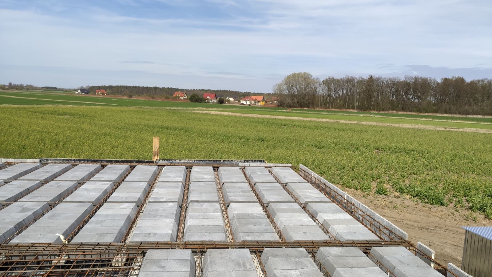 Zalany fundament z pustaków szalunkowych i zbrojenia, widok na zielone pole i zabudowania w oddali, słoneczny dzień.