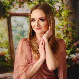 Portret uśmiechniętej blondynki w zwiewnej, różowej sukience na tle kwiatowej dekoracji okiennej, dłonie delikatnie dotykają twarzy, podkreślając makijaż.