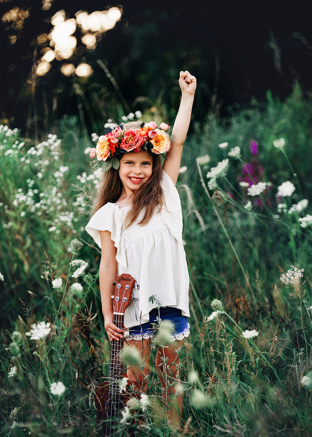 Uśmiechnięta dziewczynka w wianku z kwiatów, stojąca na łące, trzymająca ukulele i unosząca jedną rękę w górę.