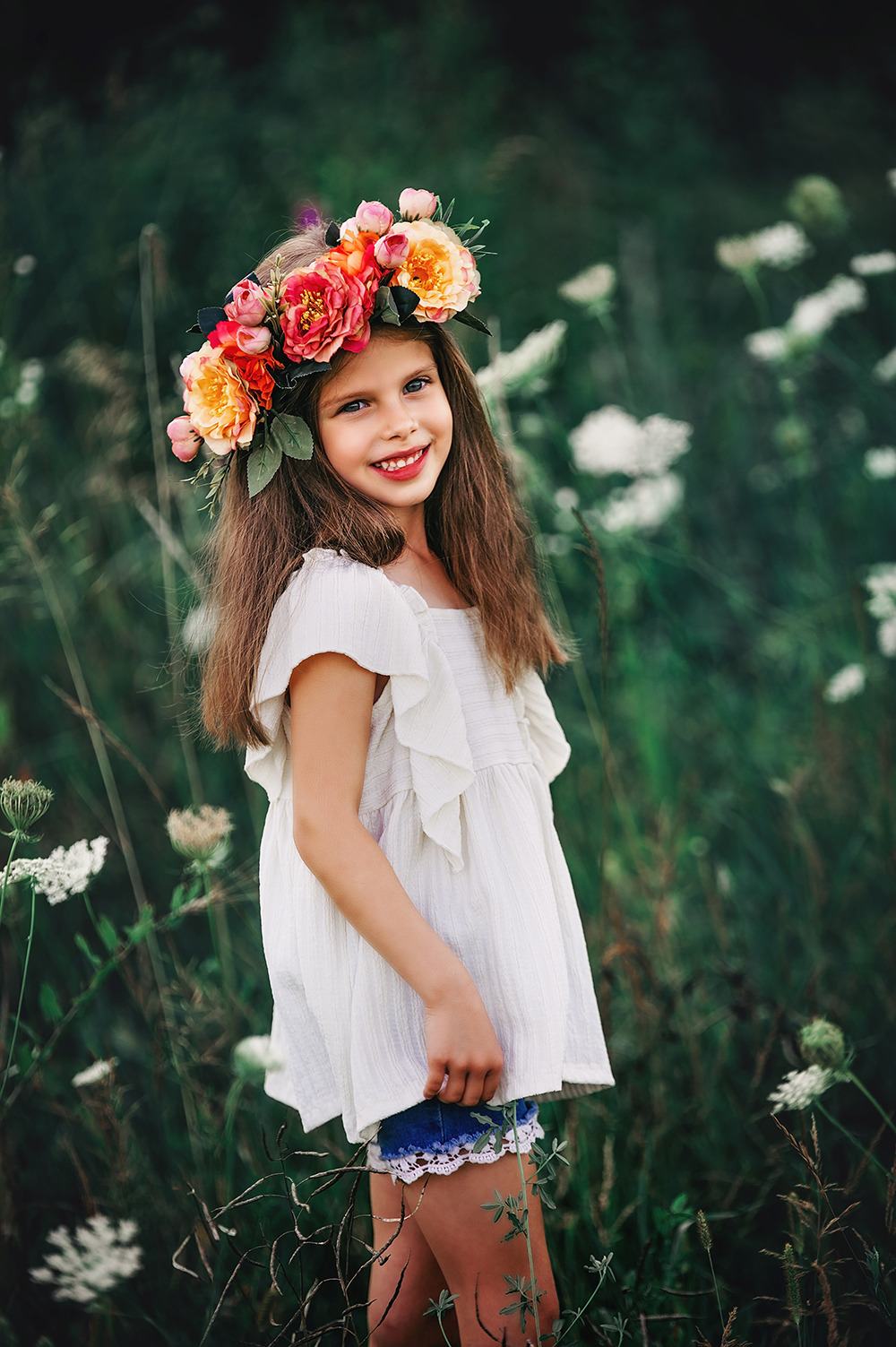 Uśmiechnięta dziewczynka z wiankiem z różowych i pomarańczowych kwiatów na głowie, ubrana w białą bluzkę i dżinsowe szorty z koronką, pozuje na łące pełnej białych kwiatów.