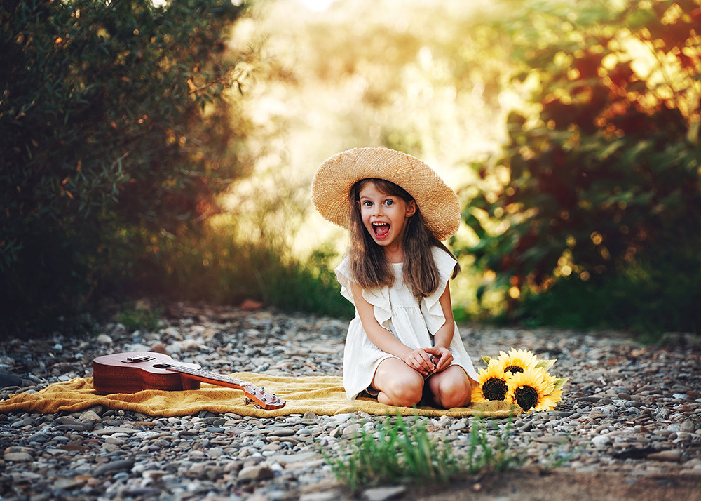 Uśmiechnięta dziewczynka w słomkowym kapeluszu siedzi na kocu z ukulele i słonecznikami na tle zieleni