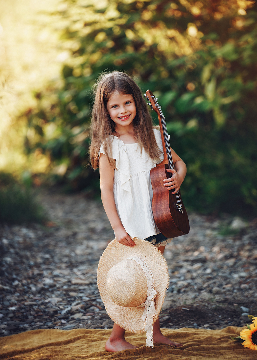 Uśmiechnięta dziewczynka z ukulele i słomkowym kapeluszem na tle zieleni.