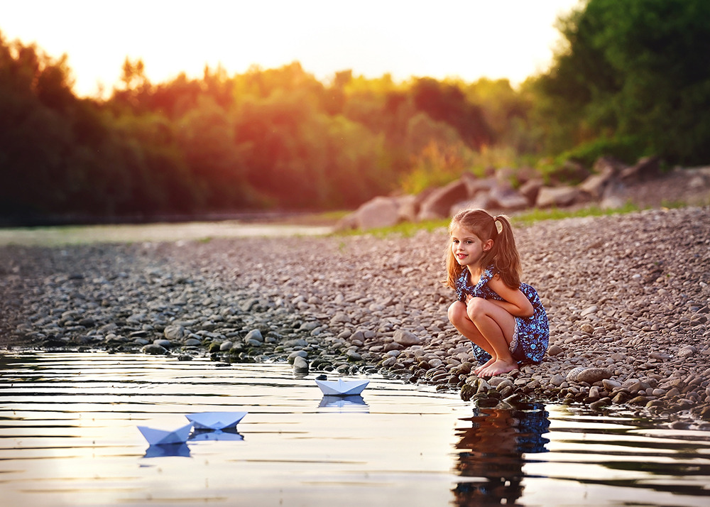 Dziewczynka w kwiecistej sukience kuca nad brzegiem rzeki, puszczając papierowe łódki na wodę o zachodzie słońca, z kamienistym brzegiem i zalesionym tłem.