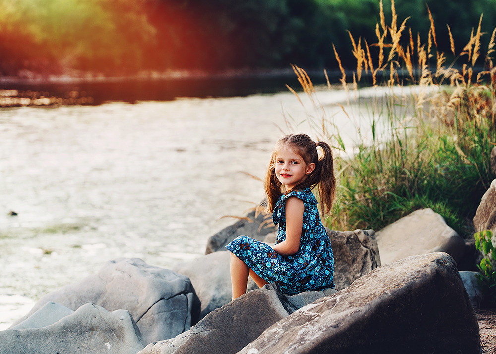 Uśmiechnięta dziewczynka w kwiecistej sukience siedzi na kamieniach nad brzegiem rzeki, w tle woda i trawy.