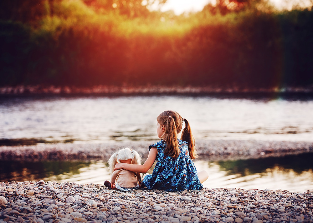 Dziewczynka w kwiecistej sukience siedzi tyłem na kamienistej plaży, obejmując lalkę z blond włosami, w tle woda i zieleń w ciepłym świetle zachodzącego słońca.