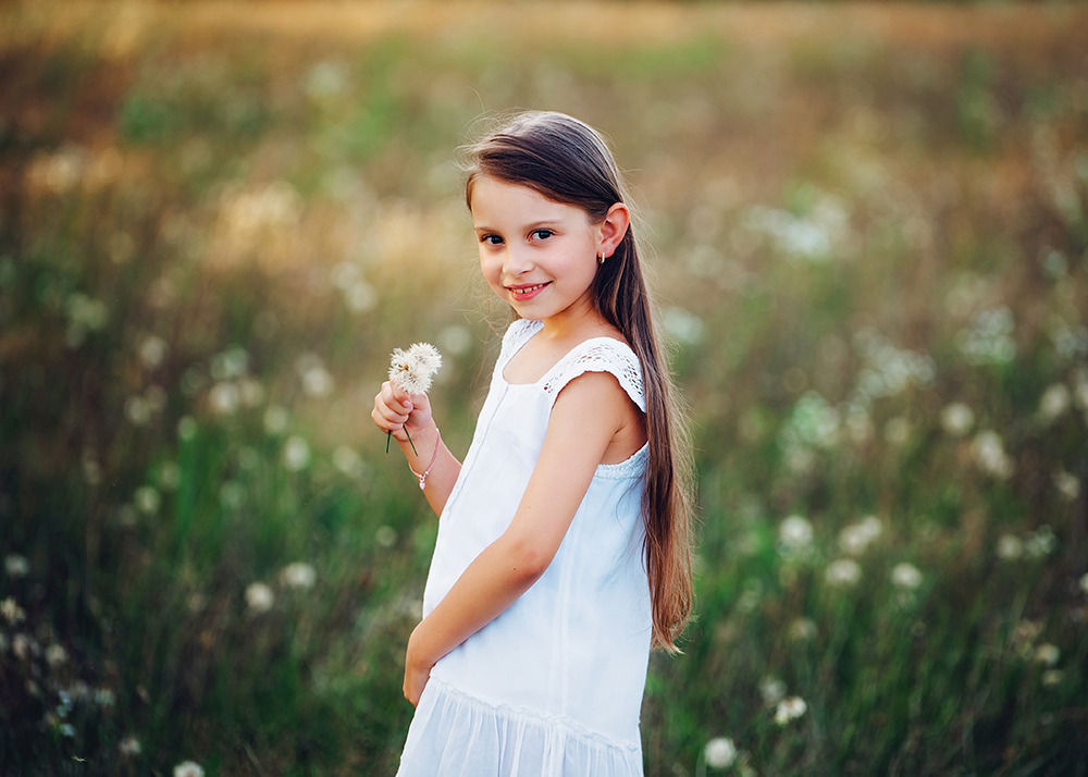 Uśmiechnięta dziewczynka w białej sukience trzyma dmuchawca na łące pełnej przekwitłych kwiatów, delikatne światło słoneczne pada na tło.