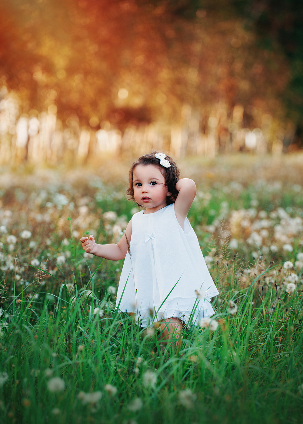 Urocza dziewczynka w białej sukience i z kokardą we włosach stoi na łące pełnej dmuchawców, z rozmarzonym spojrzeniem i ręką uniesioną do góry.